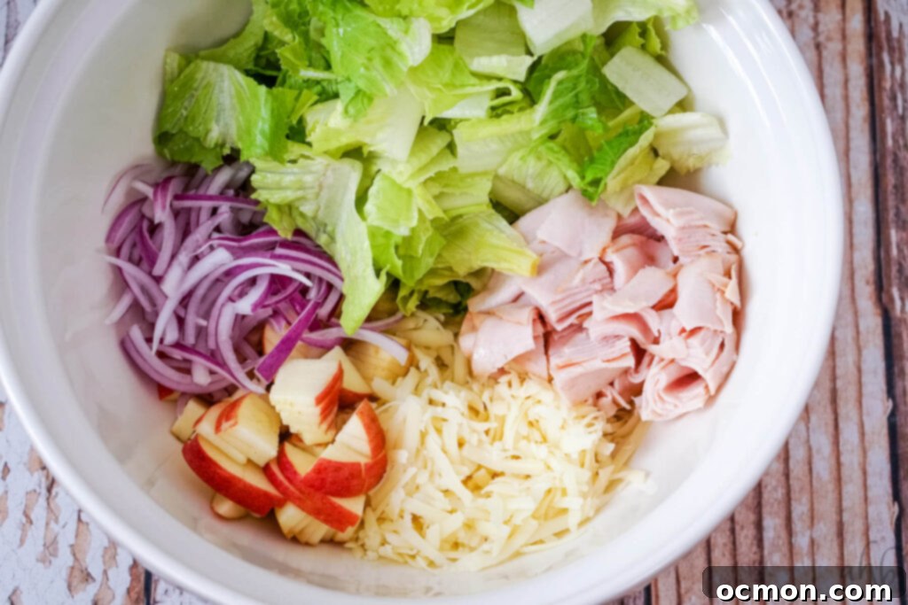 A white bowl of chopped lettuce, turkey, and apples with shredded swiss cheese and red onion.