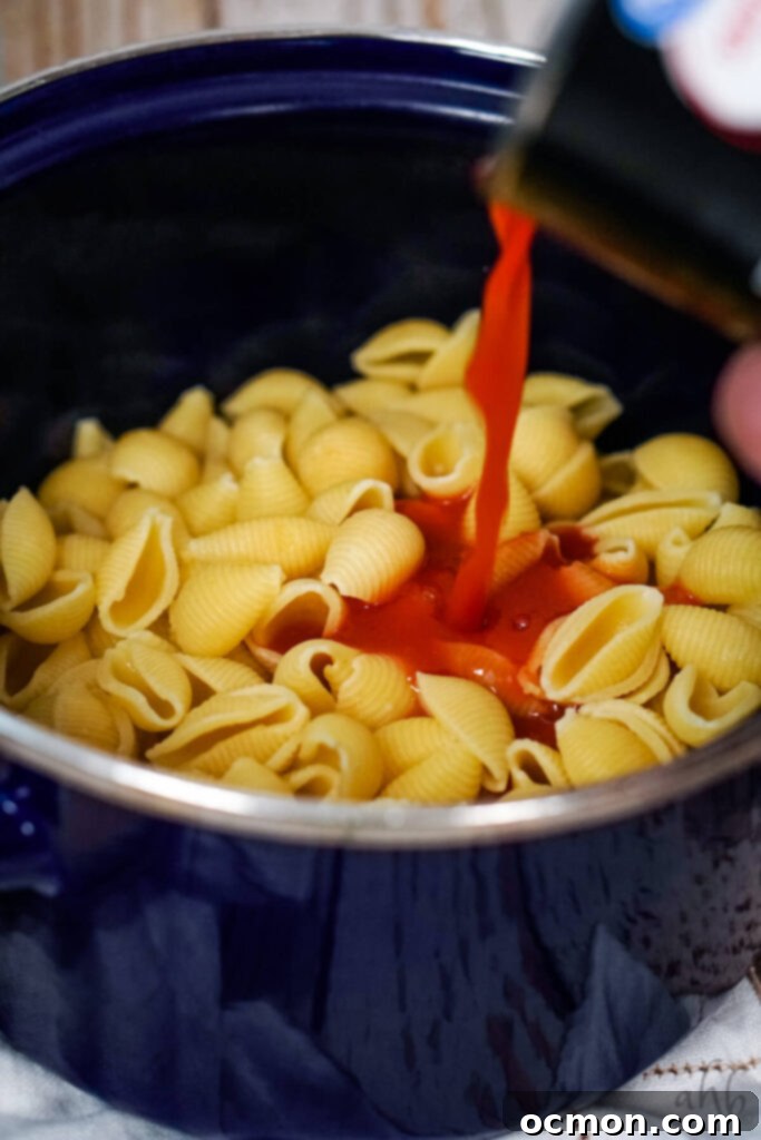Tomato juice is carefully added to the shell pasta in the blue stockpot, preparing for simmering.