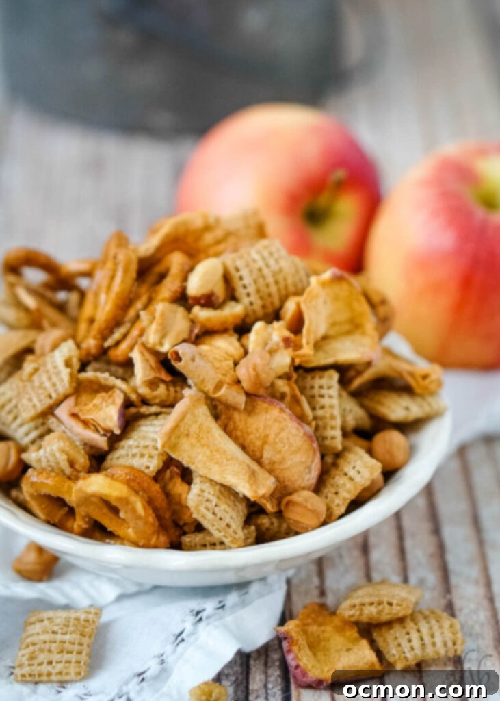 Caramel Apple Crunch Mix 3 A serving of Caramel Apple Chex Mix in a white dish, artfully arranged in front of two fresh apples.