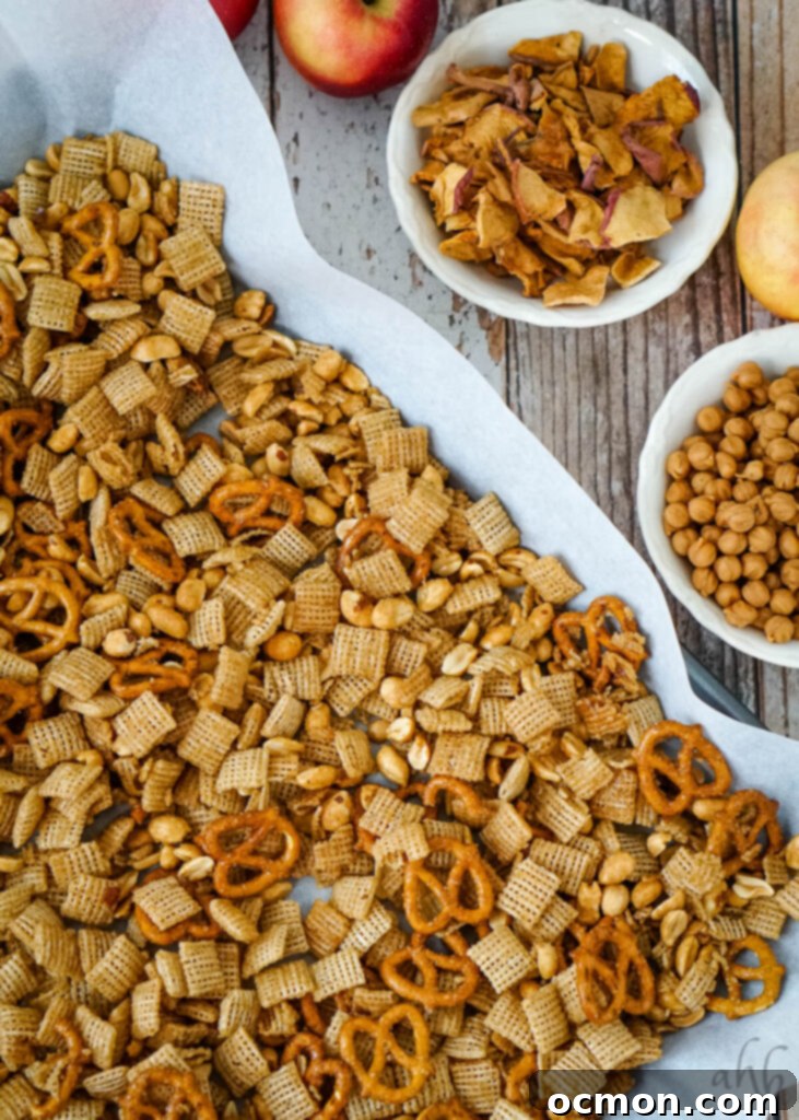 Caramel Apple Crunch Mix 8 The coated cereal mixture is spread out on a parchment-lined baking tray to cool, alongside bowls of apple chips and caramel chips, ready to be added.