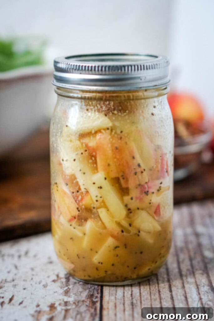 A mason jar filled with shimmering homemade maple vinaigrette, with thinly sliced apples submerged, preventing browning and infusing flavor. 