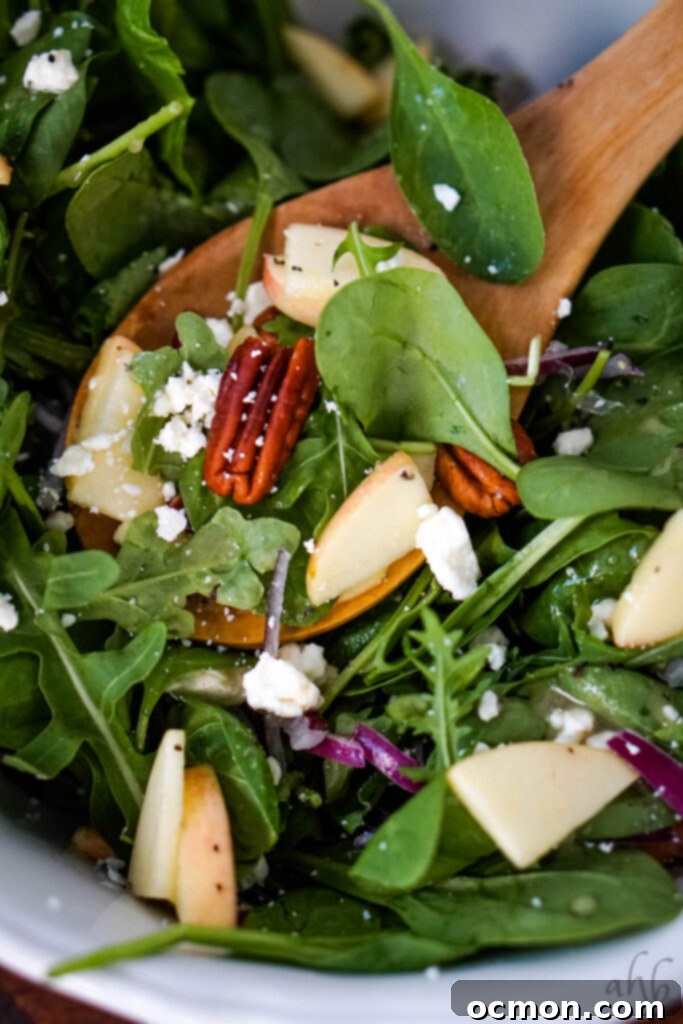 A wooden spoon carefully scooping a delightful mix of toasted pecan, fresh spinach, crisp apple slices, and creamy feta cheese from a prepared bowl of Apple Feta Spinach Salad. 