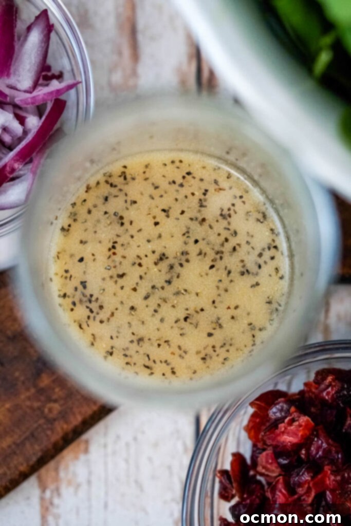 An open mason jar brimming with a perfectly emulsified homemade maple dressing, positioned next to a small bowl of ruby-red dried cranberries, ready for assembly. 
