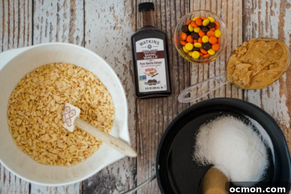 A bowl of Rice Krispies, a bottle of vanilla extract, a serving of Reese's pieces, 3/4 cup of peanut butter, and a small saucepan of corn syrup, brown sugar, and granulated sugar.