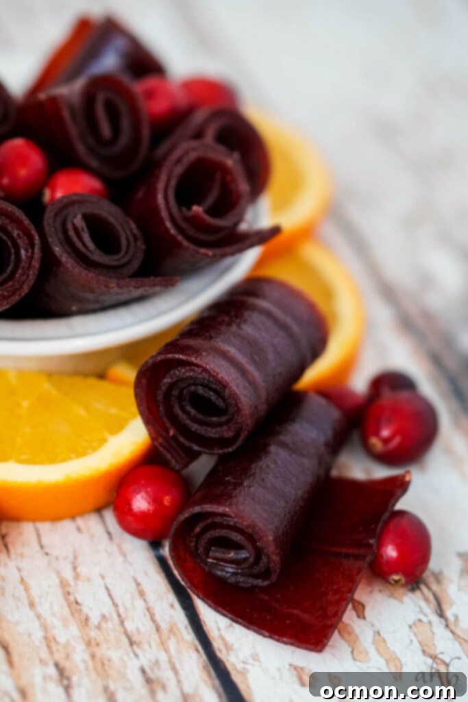 Two Cranberry orange fruit leather rolls with several cranberries and orange slices next to a bowl of cranberry orange fruit leather rolls. 