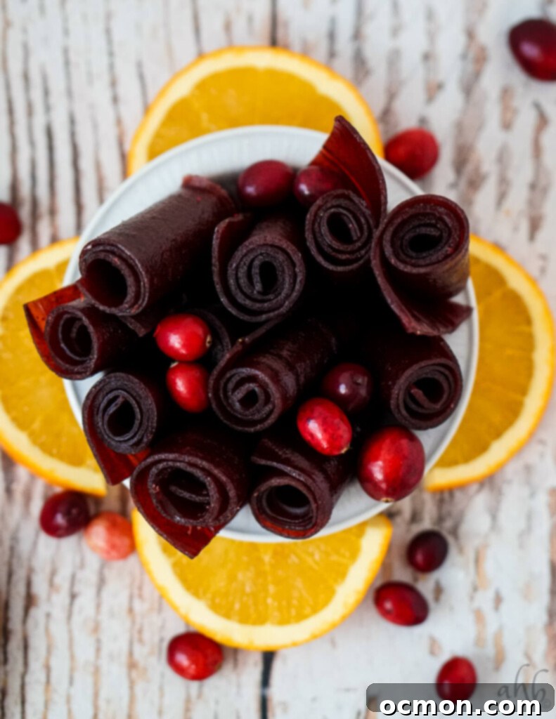 A white bowl of cranberry fruit leather rolls with orange slices and cranberries. 