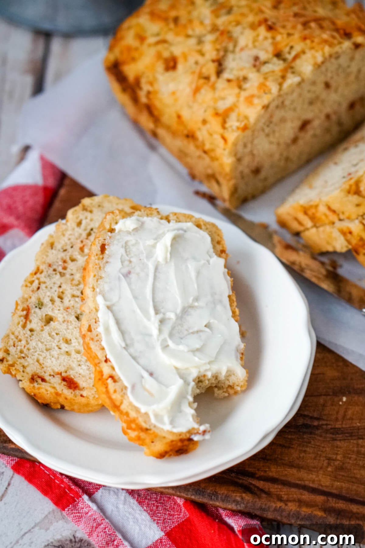A beautifully sliced loaf of Sun Dried Tomato and Parmesan Beer Bread, with two individual slices served on a white plate, one spread with cream cheese and a bite taken, showcasing the vibrant red flecks and cheesy crust.