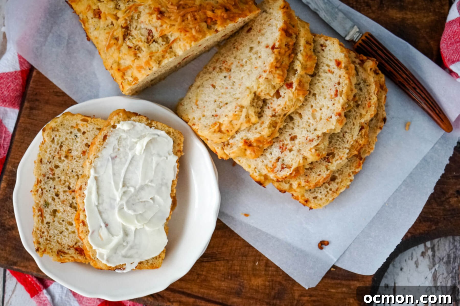 A sliced loaf of Sun Dried Tomato and Parmesan Beer Bread is presented alongside two slices on a white plate, ready to be enjoyed with cream cheese.