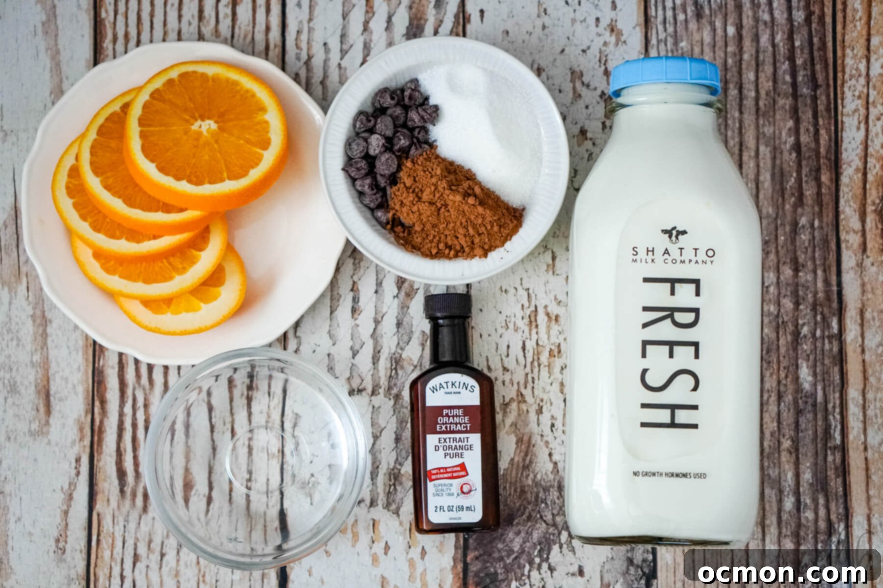 Slices of orange on a white plate, a bowls of granulated sugar, cocoa powder, and semi-sweet chocolate chips in a white bowl, a bowl of water, a bottle of orange extract, and a bottle of Shatto milk.