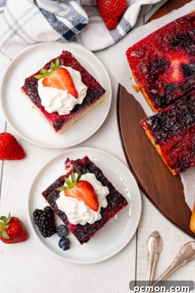 Two servings of Berry Upside down cake are served on white plates with whipped cream and strawberries next to the serving board with the rest of the cake.