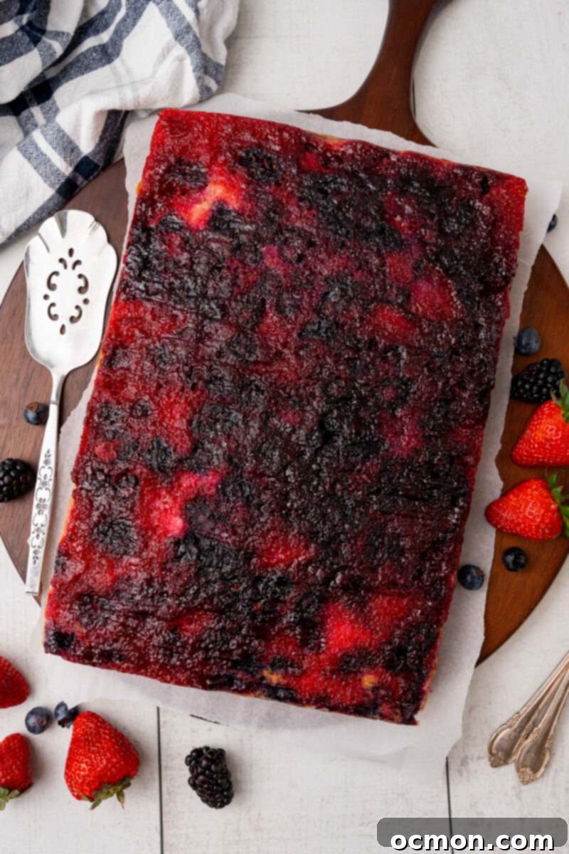 A cooled Berry Upside Down Cake sits on parchment on a serving platter surrounded by berries.
