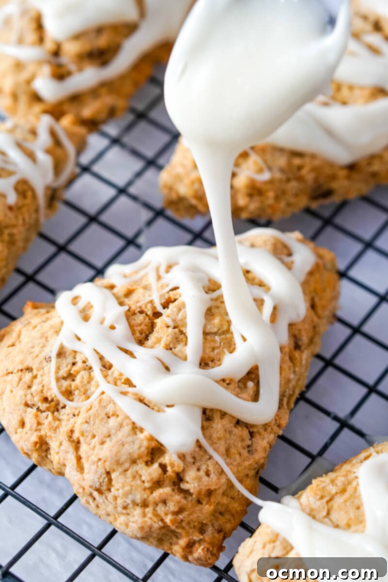 A delicate cream cheese glaze is being artfully drizzled over a batch of freshly baked and cooled carrot cake mix scones, arranged neatly on a wire cooling rack.