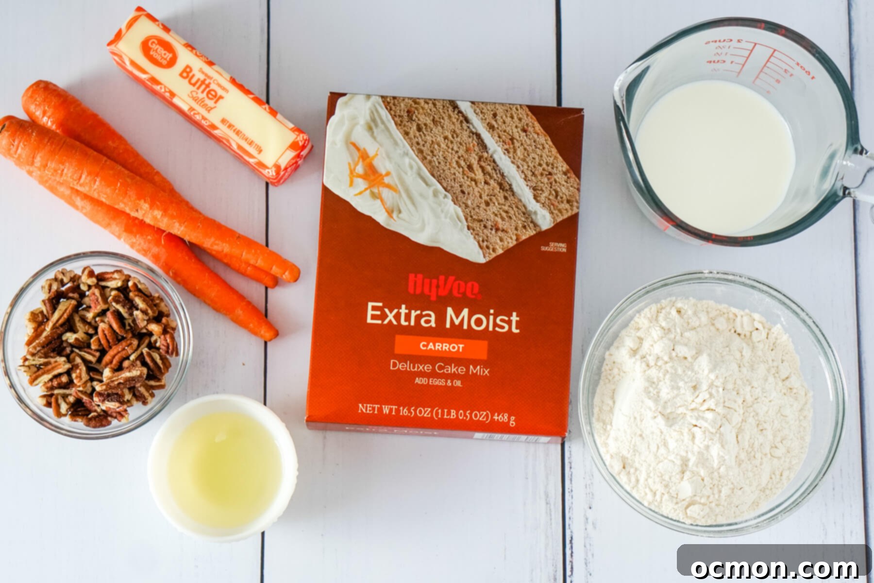 A selection of fresh ingredients laid out: pecan pieces, three whole carrots, an egg, a stick of cold butter, a measuring cup of milk, a box of carrot cake mix, and a bowl of all-purpose flour.