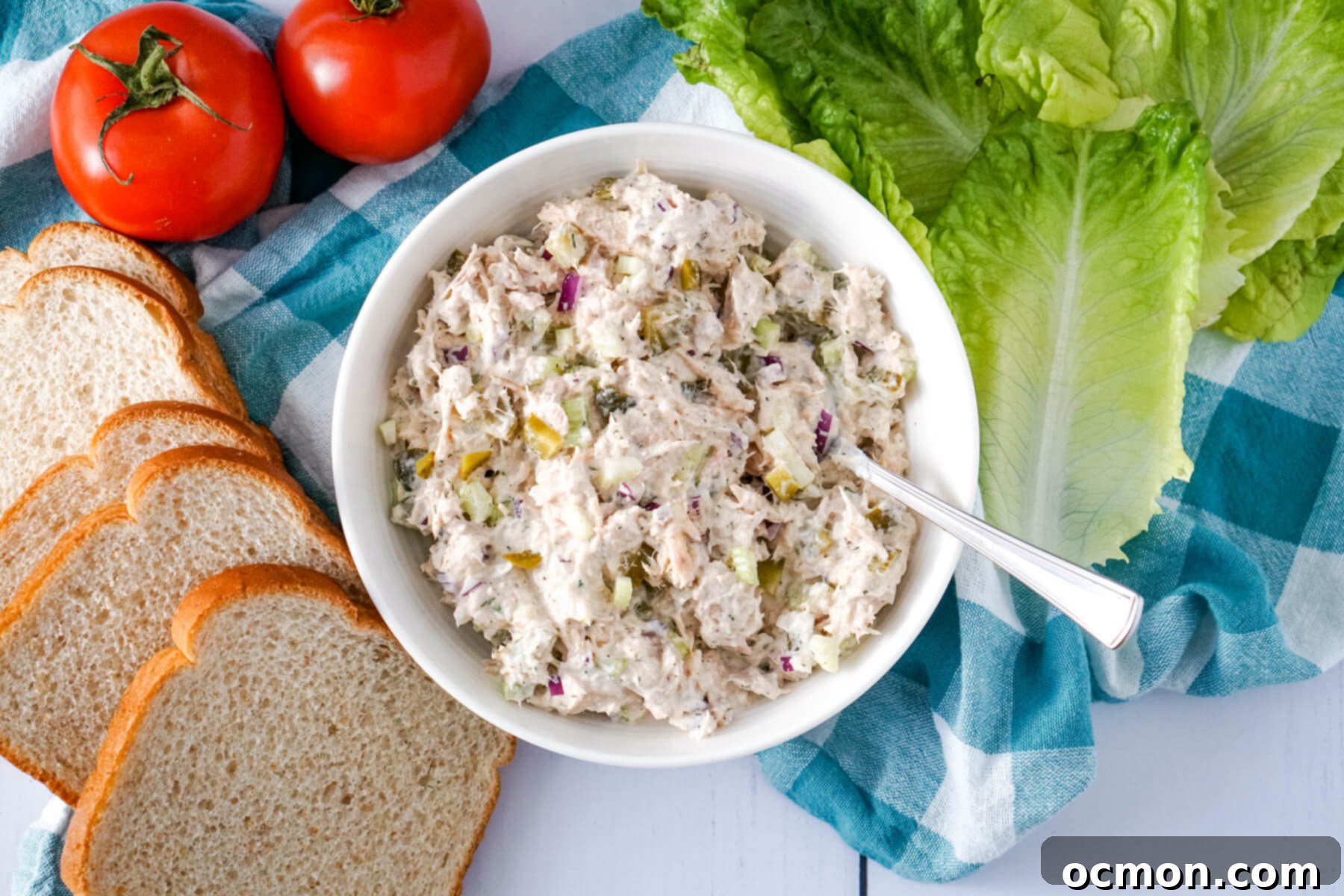 Tangy Dill Pickle Tuna Salad 3 A white bowl of Dill Pickle Tuna Salad sits next to lettuce leaves, two whole tomatoes and slices of bread.