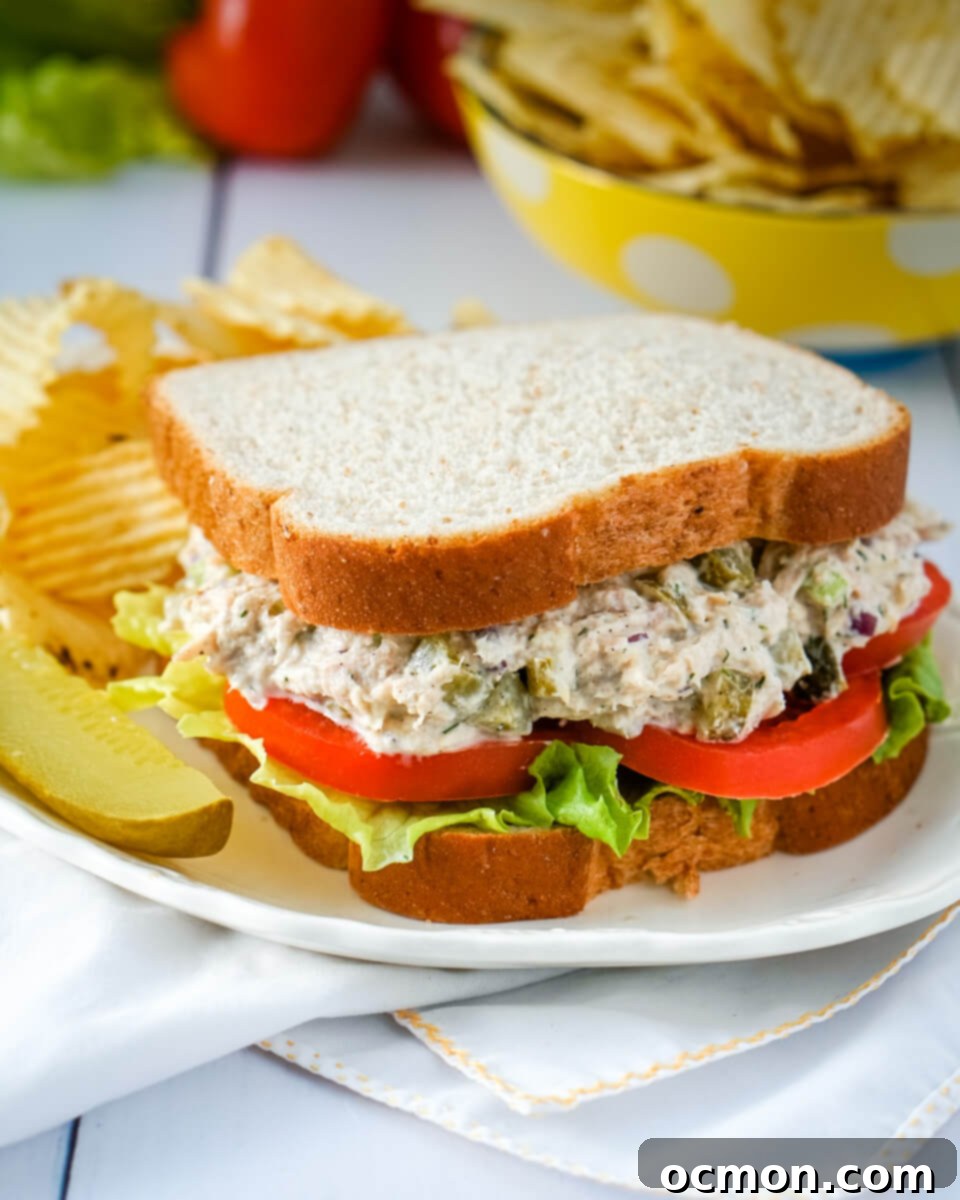 Tangy Dill Pickle Tuna Salad 6 A Dill Pickle Tuna Salad Sandwich is served on a white plate with a pickle and potato chips.