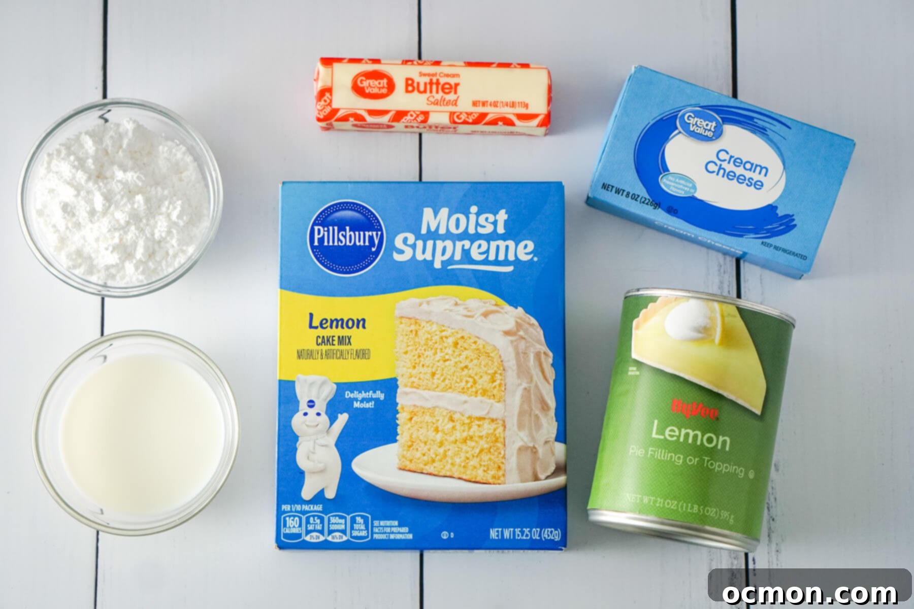Bright Lemon Dump Cake 4 Key ingredients for lemon dump cake are neatly arranged: powdered sugar, milk, a box of lemon cake mix, a stick of butter, a block of cream cheese, and a can of lemon pie filling.