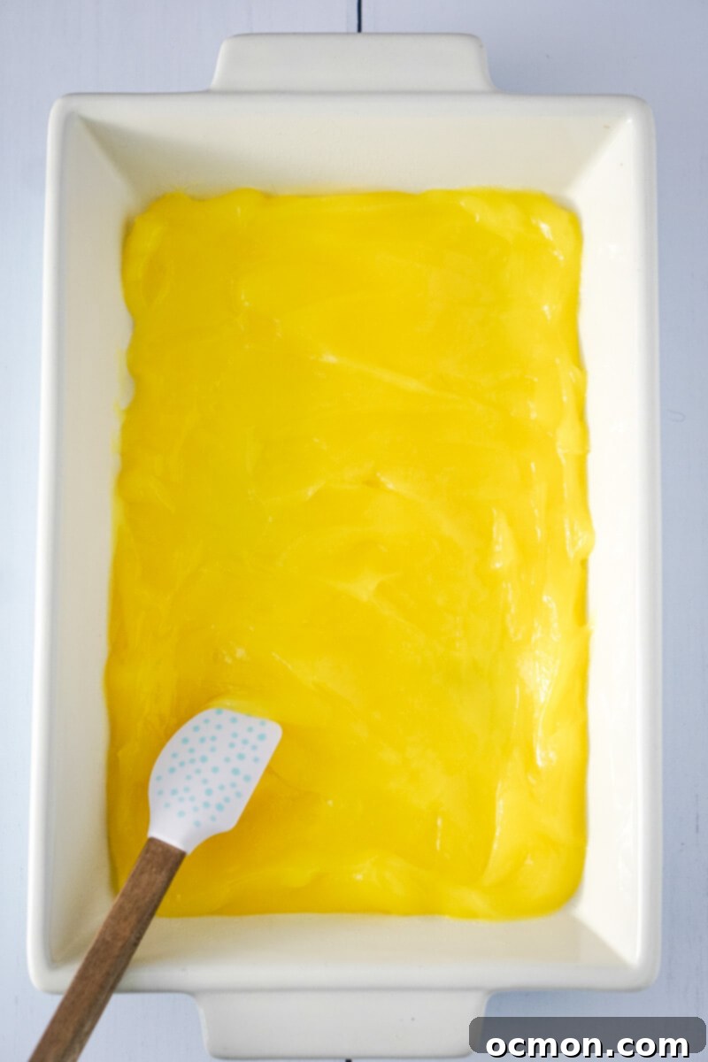 Bright Lemon Dump Cake 5 The bright yellow lemon pie filling is meticulously spread into an even layer across the bottom of a rectangular baking pan, forming the base of the cake.