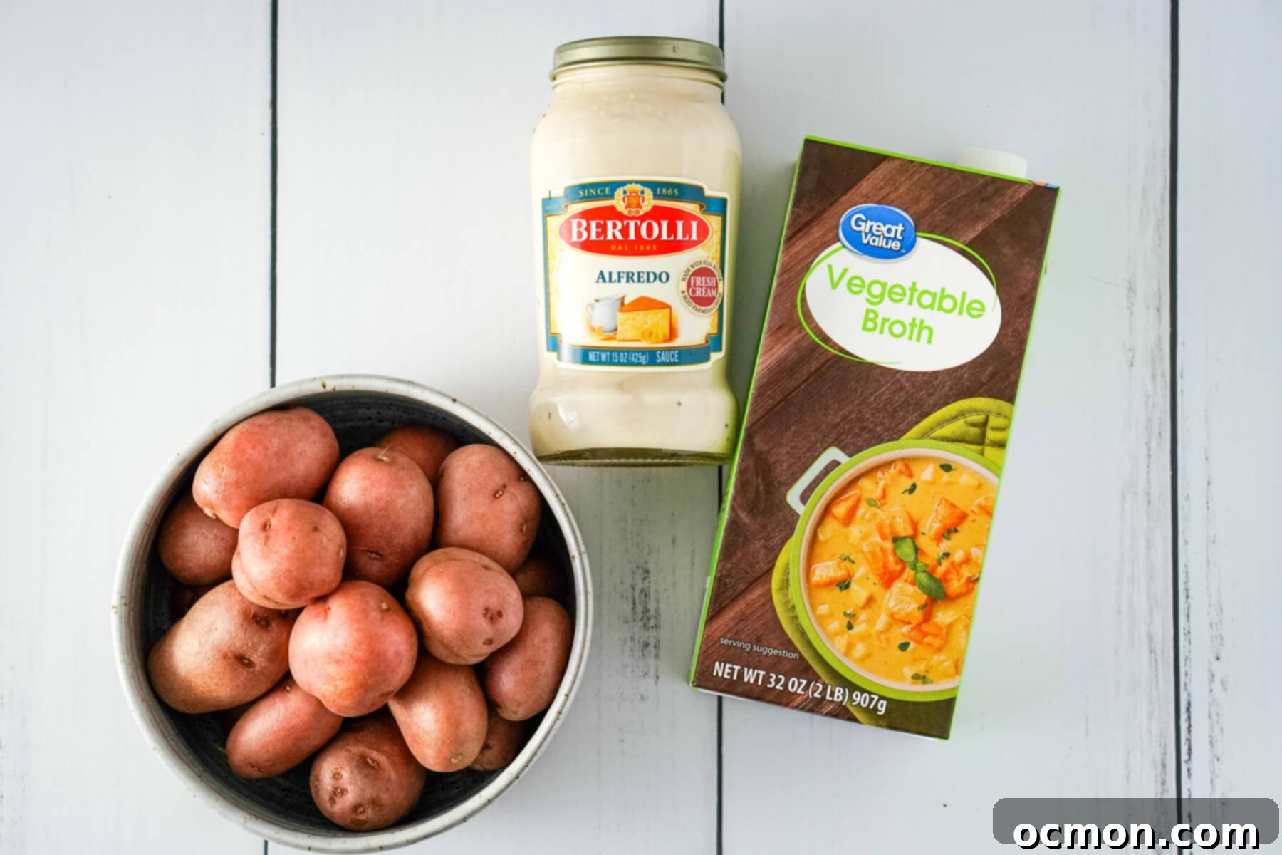 A trio of essential ingredients for potato soup: a bowl of baby red potatoes, a jar of Bertolli Alfredo sauce, and a carton of vegetable broth.