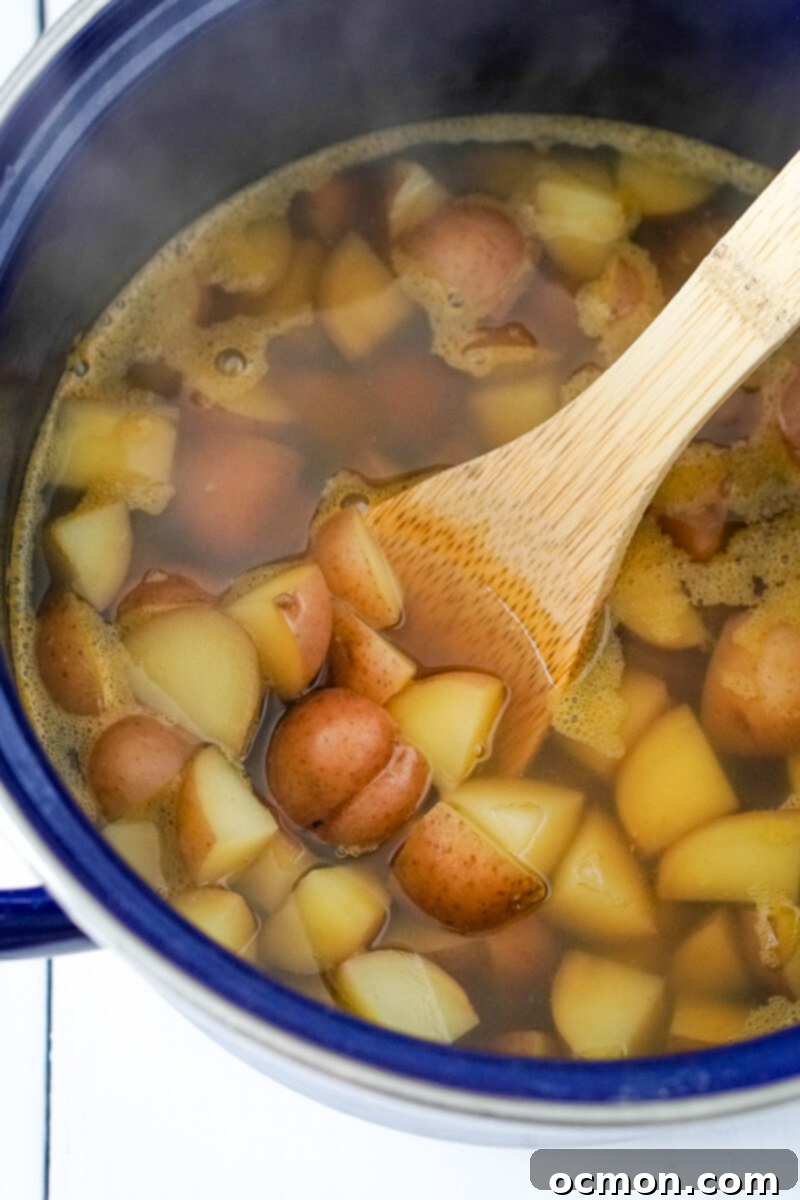 A large stock pot filled with vegetable broth and bite-sized pieces of baby red potatoes is gently stirred with a wooden spoon as it cooks.