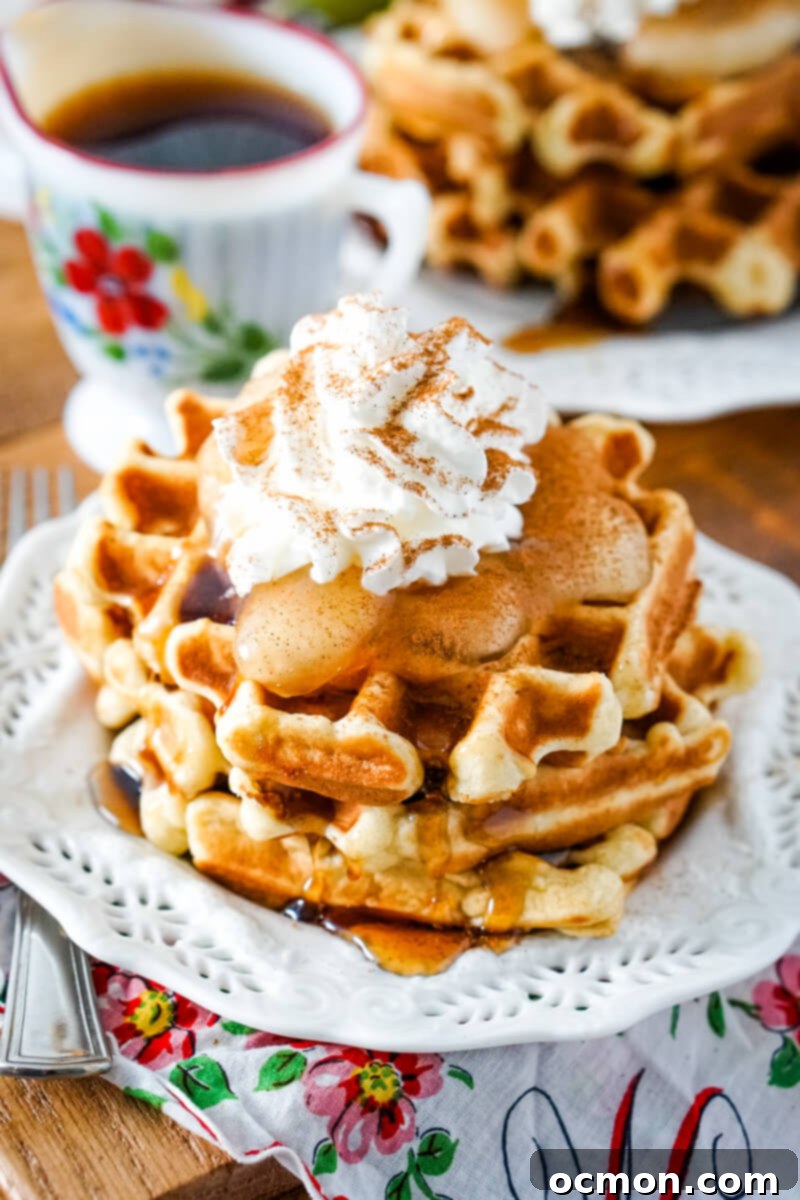 Two apple waffles are served on a white plate and topped with apple pie filling, whipped cream and cinnamon.