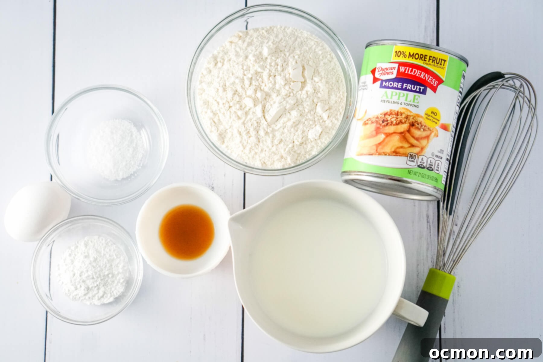 Flour, baking powder, salt, milk, and egg, vanilla extract, and a can of apple pie filling sit next to a whisk.
