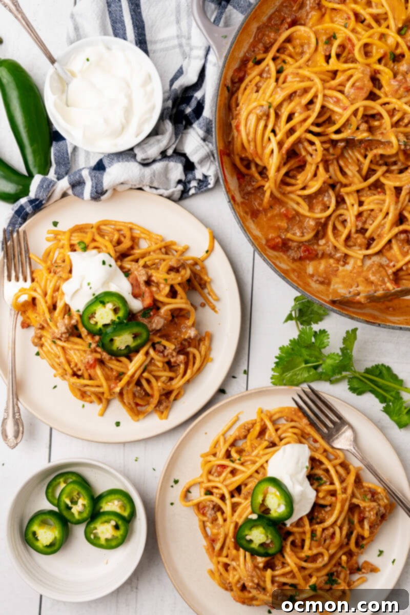 Two generous servings of Cheesy Taco Spaghetti, perfectly garnished with fresh toppings, sit on white plates alongside the large pot of creamy, cheesy taco spaghetti, ready to be enjoyed.
