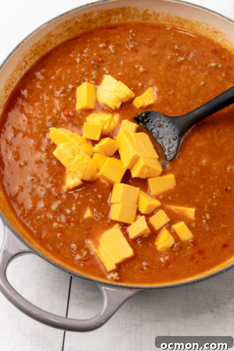 Chunks of Velveeta cheese are carefully added to the simmering sauce in the skillet, then stirred until they are completely melted and create a smooth, creamy texture.