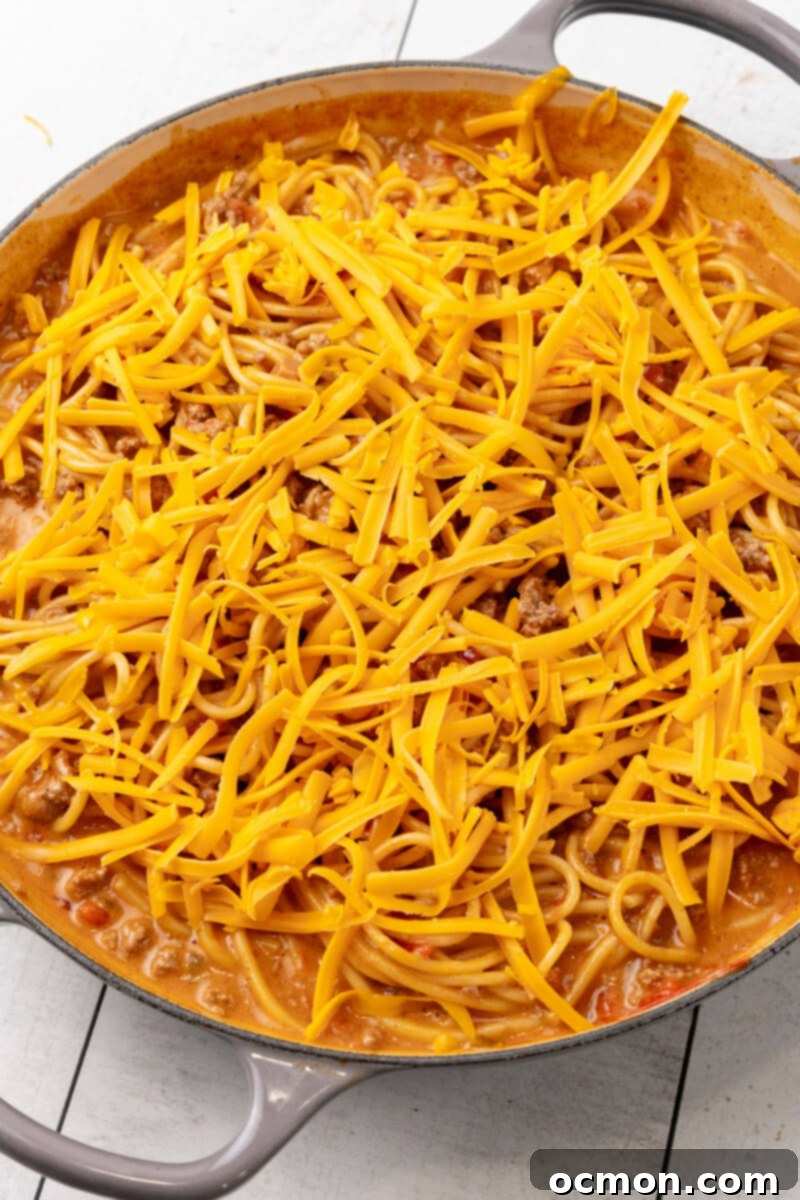A generous layer of shredded cheddar cheese is evenly added on top of the cheesy taco spaghetti in the skillet, preparing for the final melt.