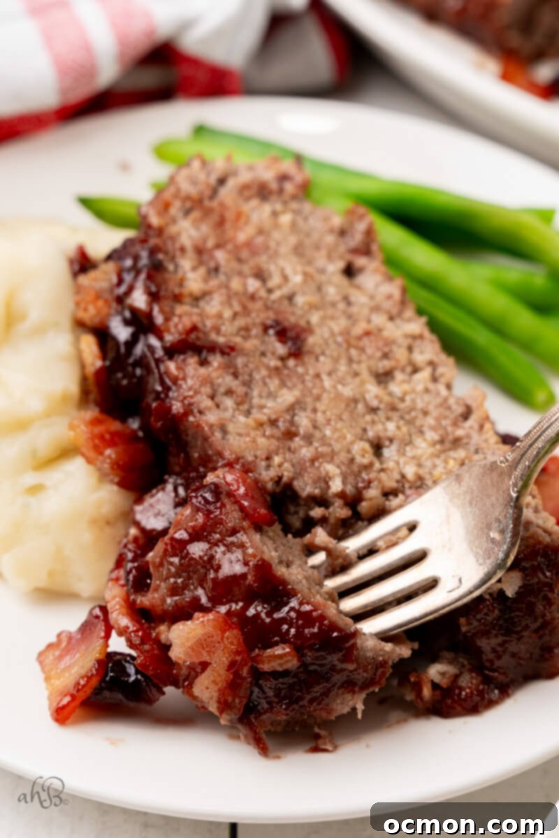 A delectable slice of Bourbon Bacon Meatloaf served alongside creamy mashed potatoes and vibrant green beans, with a fork poised for a bite.