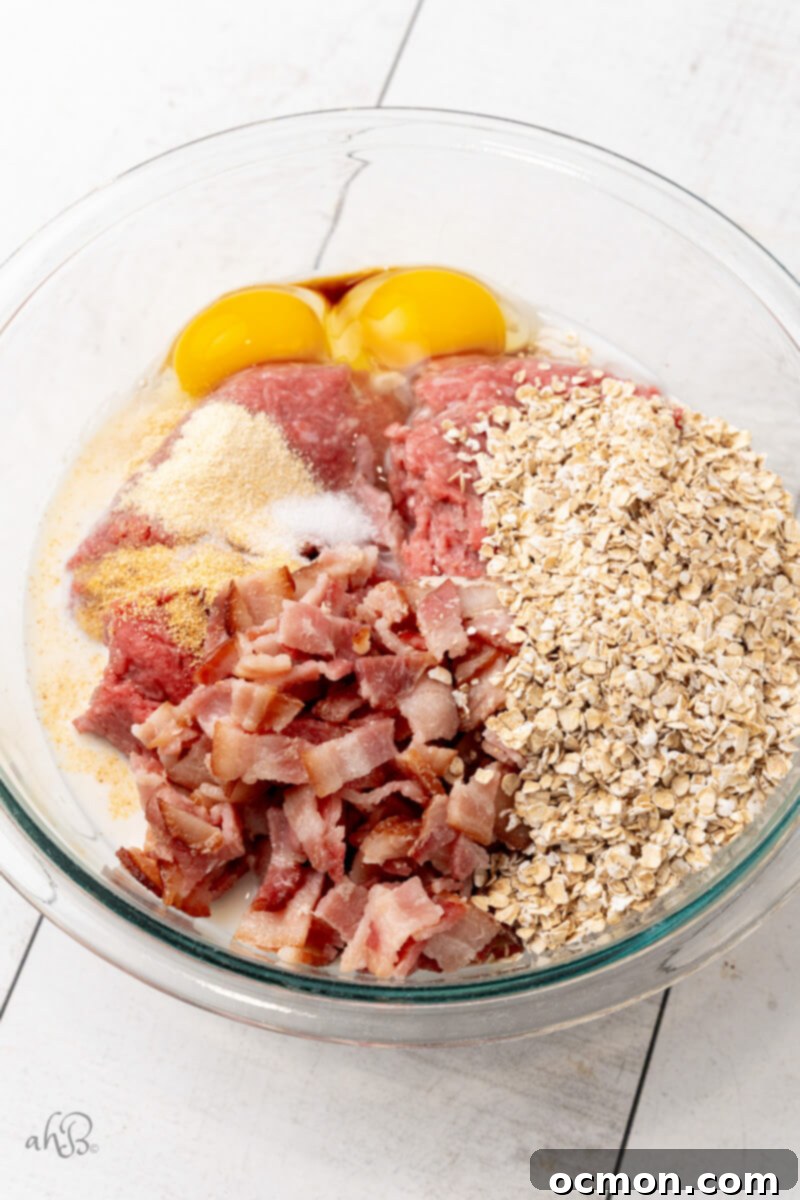 All the meatloaf ingredients, including ground beef, oats, spices, and crumbled bacon, are added to a large glass mixing bowl, ready to be combined.