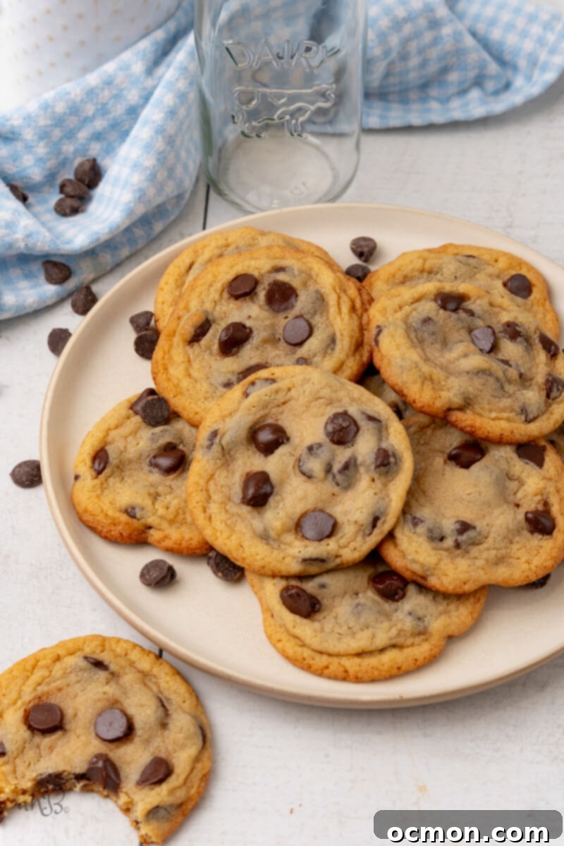 A full plate of golden-brown Malted Chocolate Chip Cookies sits next to a single cookie with a bite taken out, revealing its soft interior and melted chocolate.