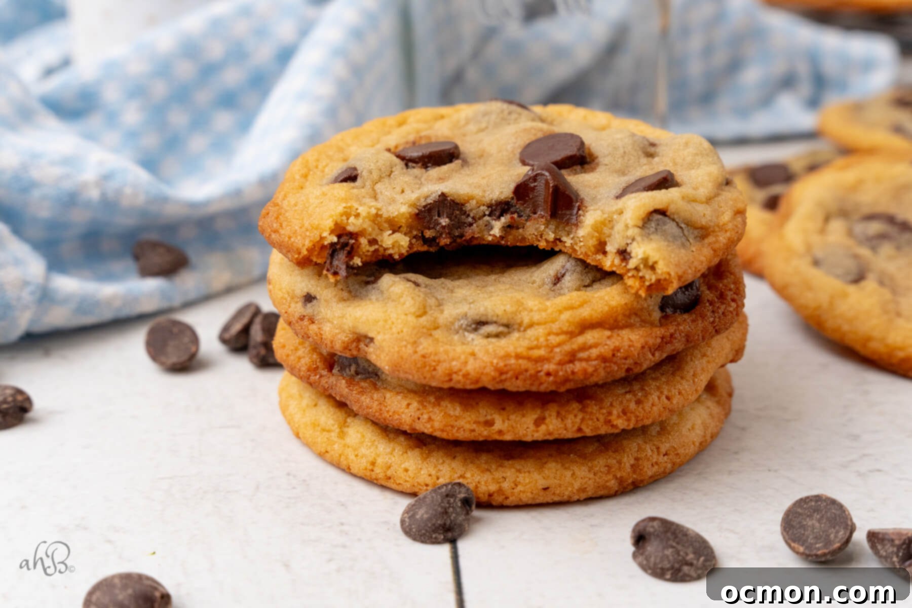 Four golden-brown Malted Milk Chocolate Chip Cookies are stacked in a tower, with scattered chocolate chips around, and the top cookie has a delicious bite taken out of it.