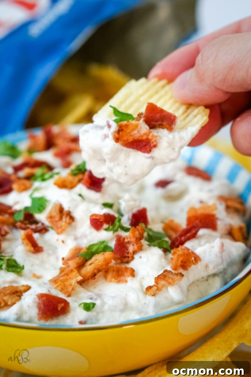A chip scoops up bacon horseradish dip from a blue and white striped bowl, highlighting its creamy texture and bacon bits.