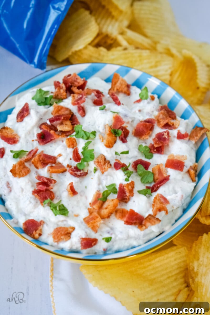 A bowl of bacon horseradish dip is served in a blue and white striped bowl and surrounded by an inviting spread of crispy potato chips.