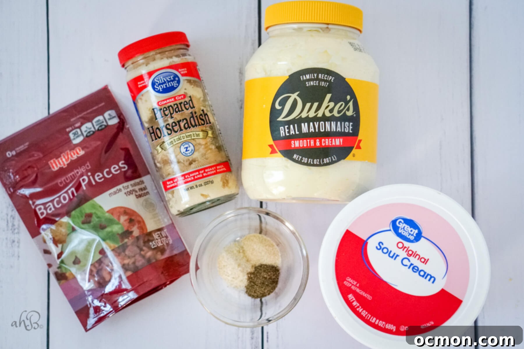 A package of real bacon pieces, prepared horseradish, a small dish of seasonings, real mayonnaise, and a container of sour cream are neatly arranged on a table, ready for mixing.