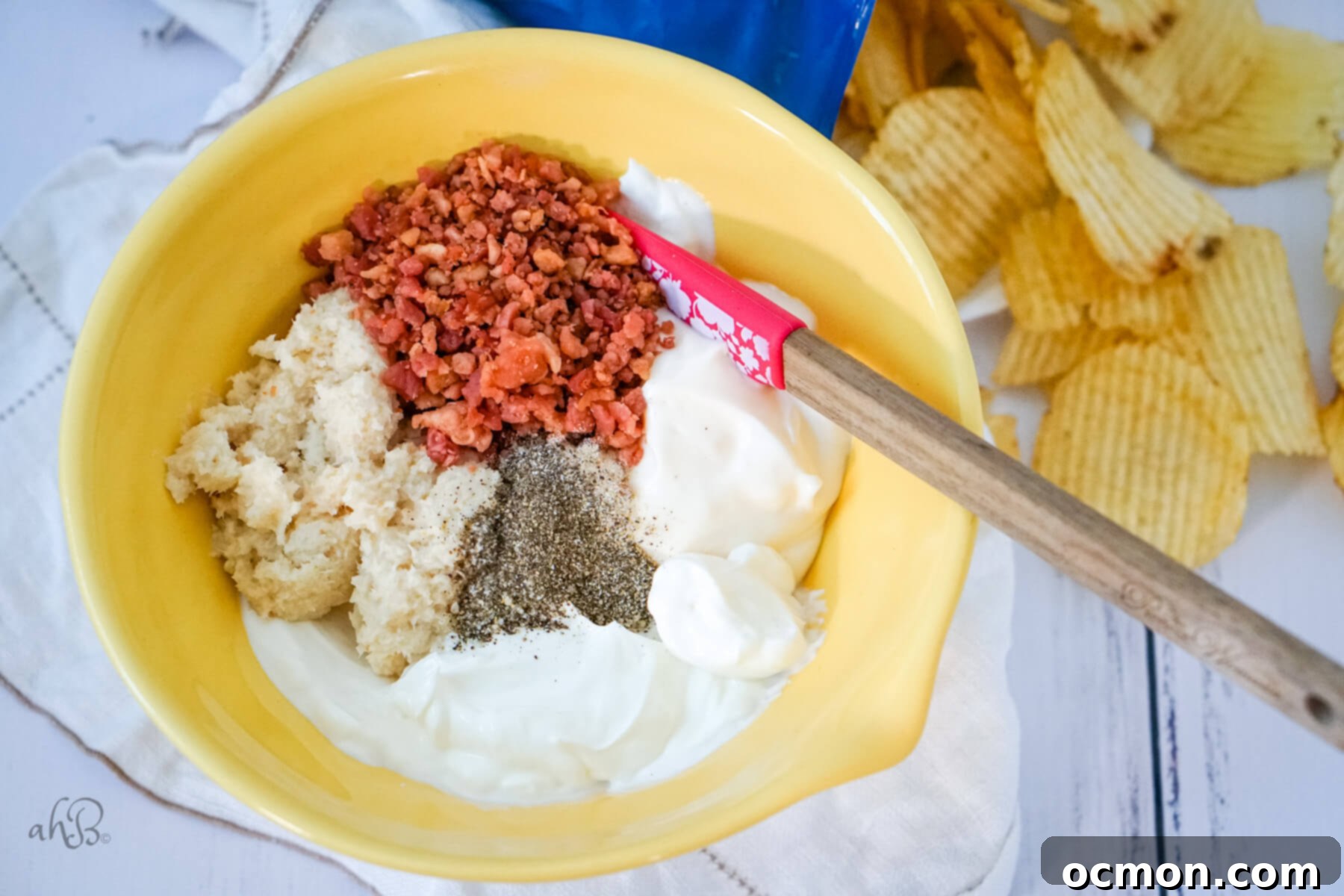 All the ingredients for the bacon horseradish dip are added to a yellow mixing bowl and are being stirred with a spatula until well combined.