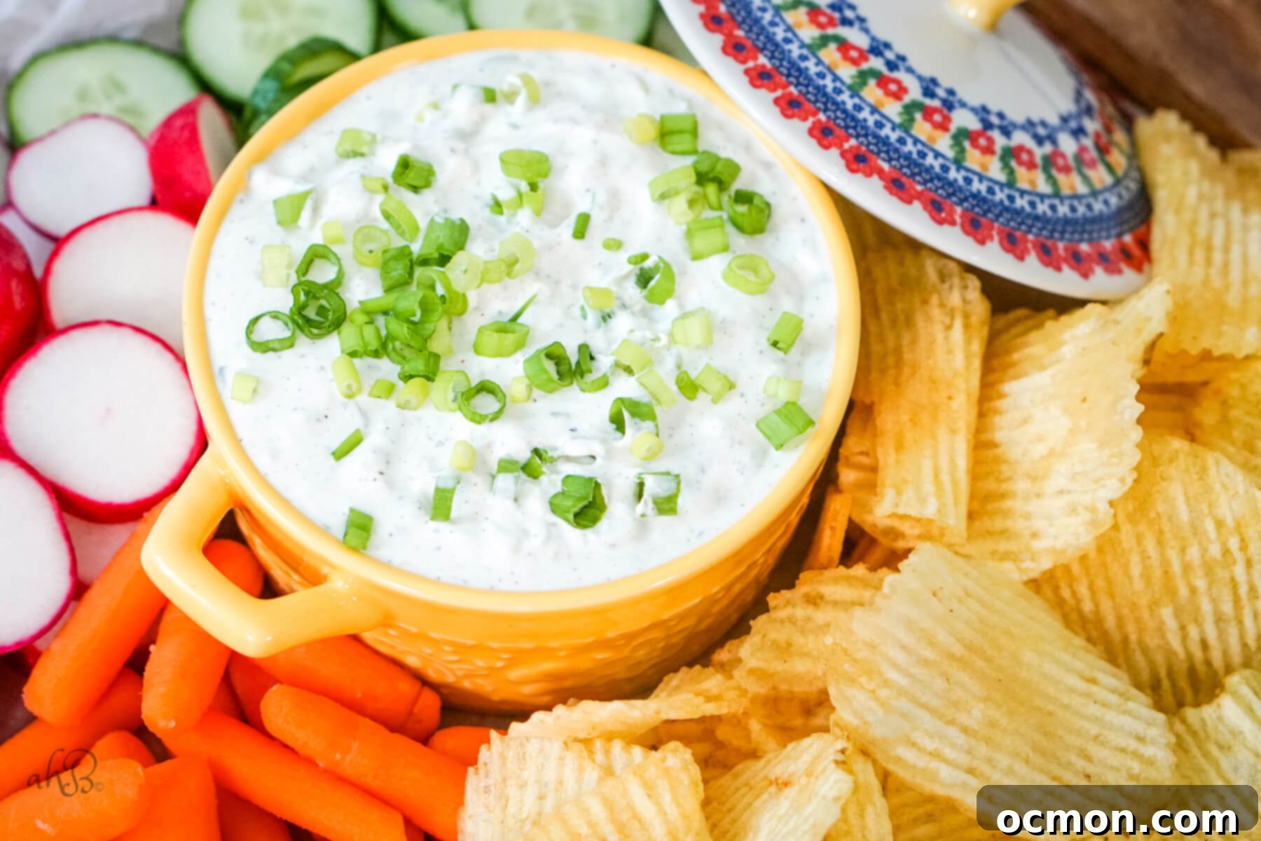 A vibrant yellow bowl of green onion dip surrounded by an assortment of crunchy potato chips and fresh-cut vegetables like carrots, cucumbers, and celery, with a lid placed beside it, ready for serving.