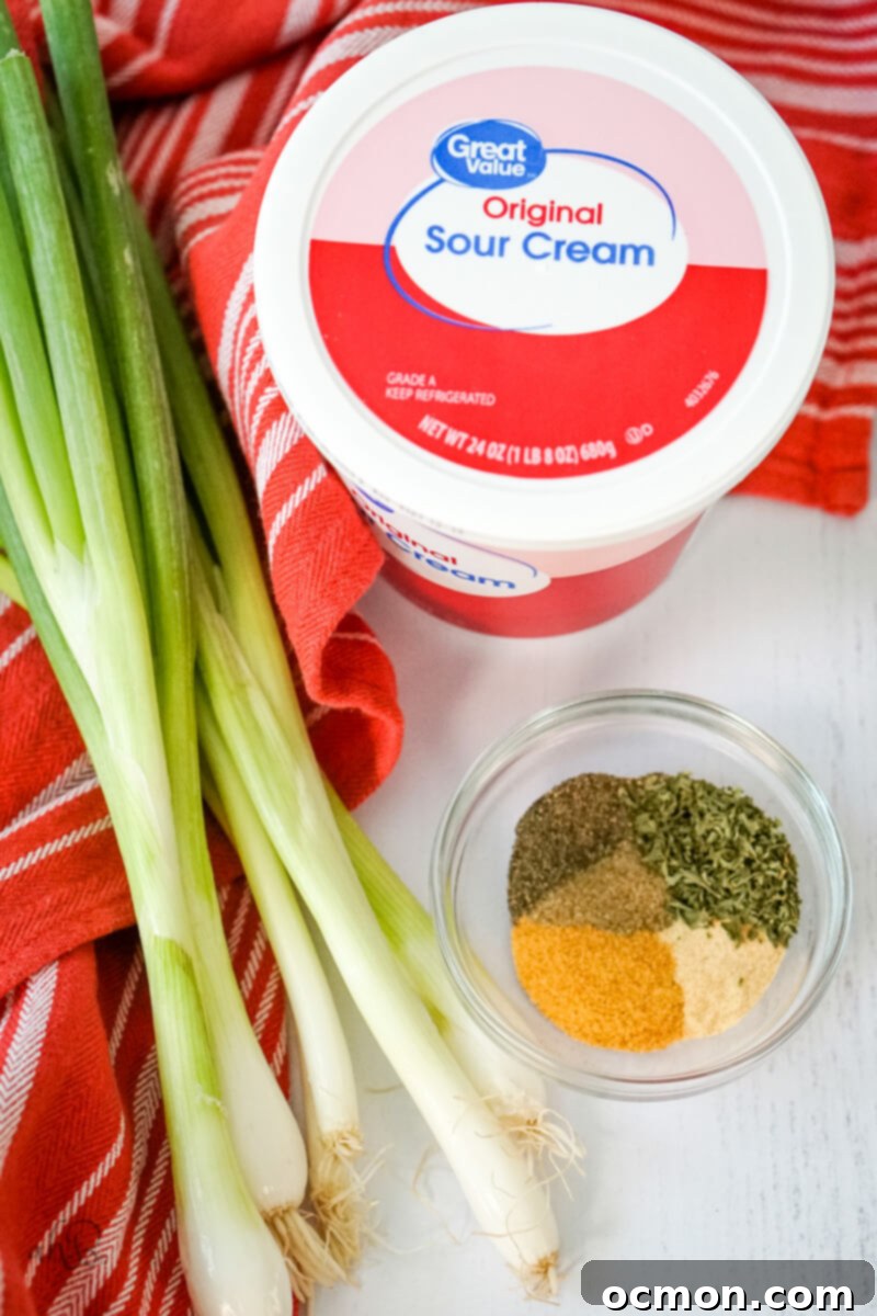 A neatly arranged flat lay of green onion dip ingredients: five vibrant green onions, a small bowl of various spices, and a large container of creamy sour cream, all resting on a rustic wooden table with a kitchen towel.