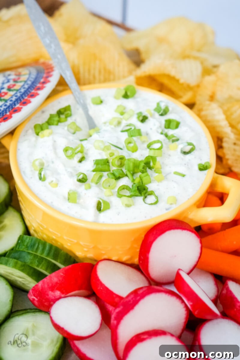 A vibrant yellow bowl of freshly made green onion dip, garnished with extra finely sliced green onions, is elegantly presented amidst a spread of potato chips and various fresh vegetables, ready to be enjoyed.