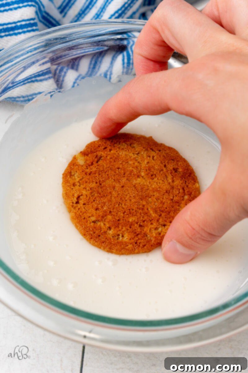 Iced Oatmeal Cookies-9 A baked oatmeal cookie is carefully dipped into a clear glass bowl of smooth, white icing, ensuring an even coating.
