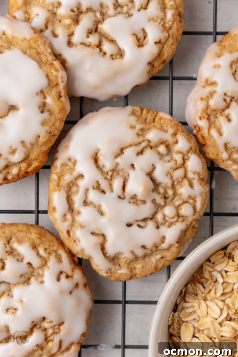 Iced Oatmeal Cookies-12 A single iced oatmeal cookie sits on a cooling rack, with more cookies and a bowl of old-fashioned oats visible in the background, showcasing the key ingredient.