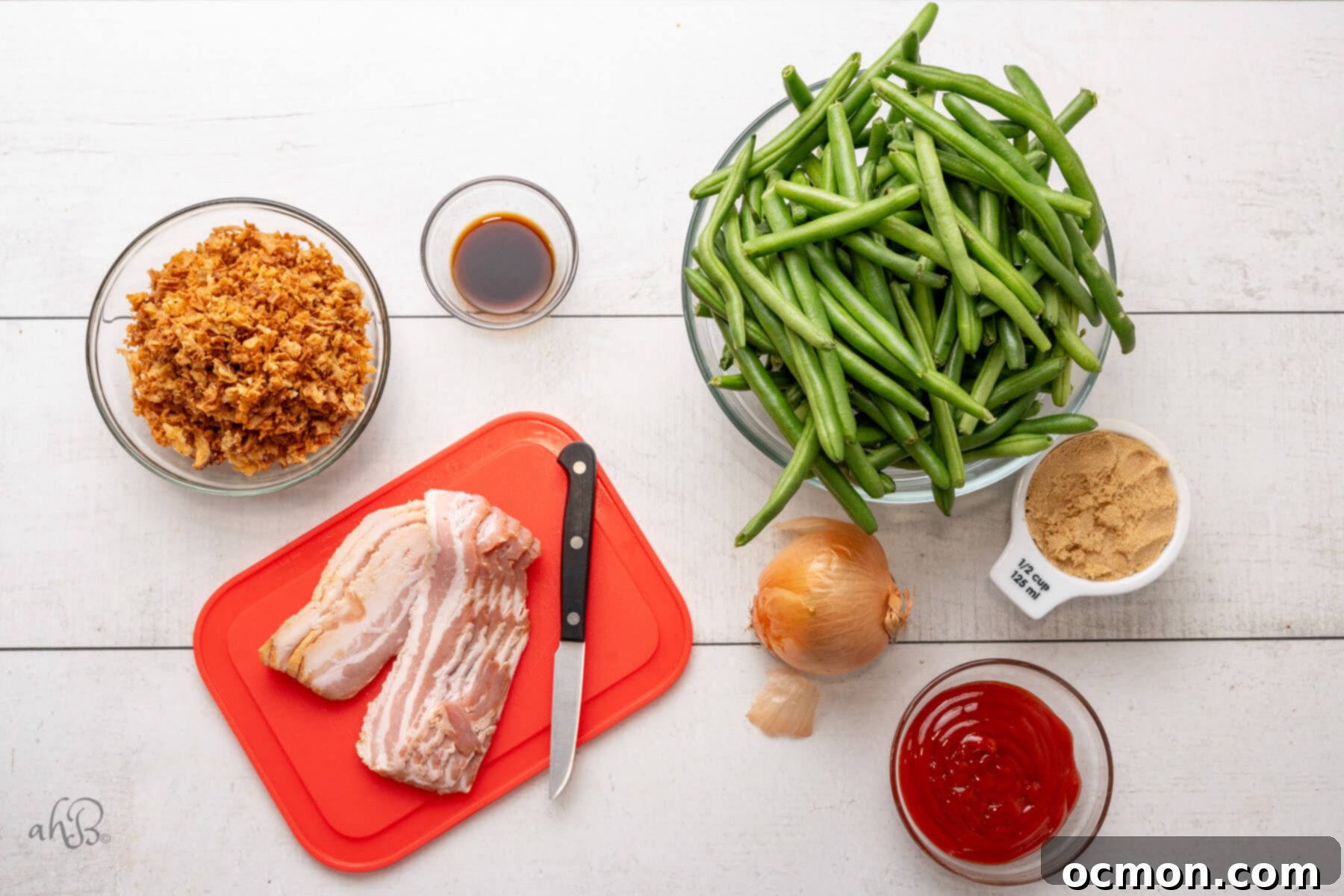 Slices of uncooked bacon, ketchup, an onion, brown sugar, green beans, Worcestershire sauce, and French fried onions laid out on a wooden surface.
