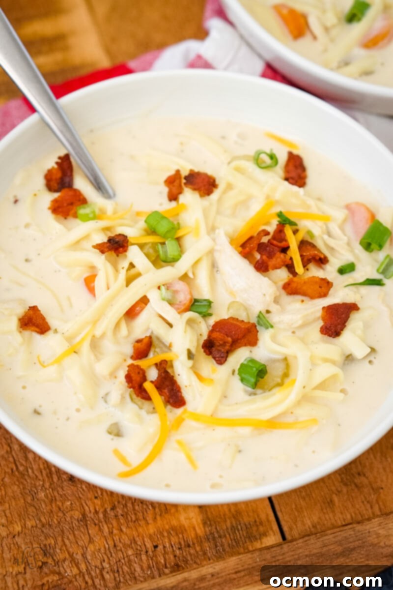 A white bowl of creamy Crack Chicken Noodle Soup topped with crispy bacon pieces, fresh chives, and a sprinkle of shredded cheese, ready to be enjoyed as a hearty comfort meal.