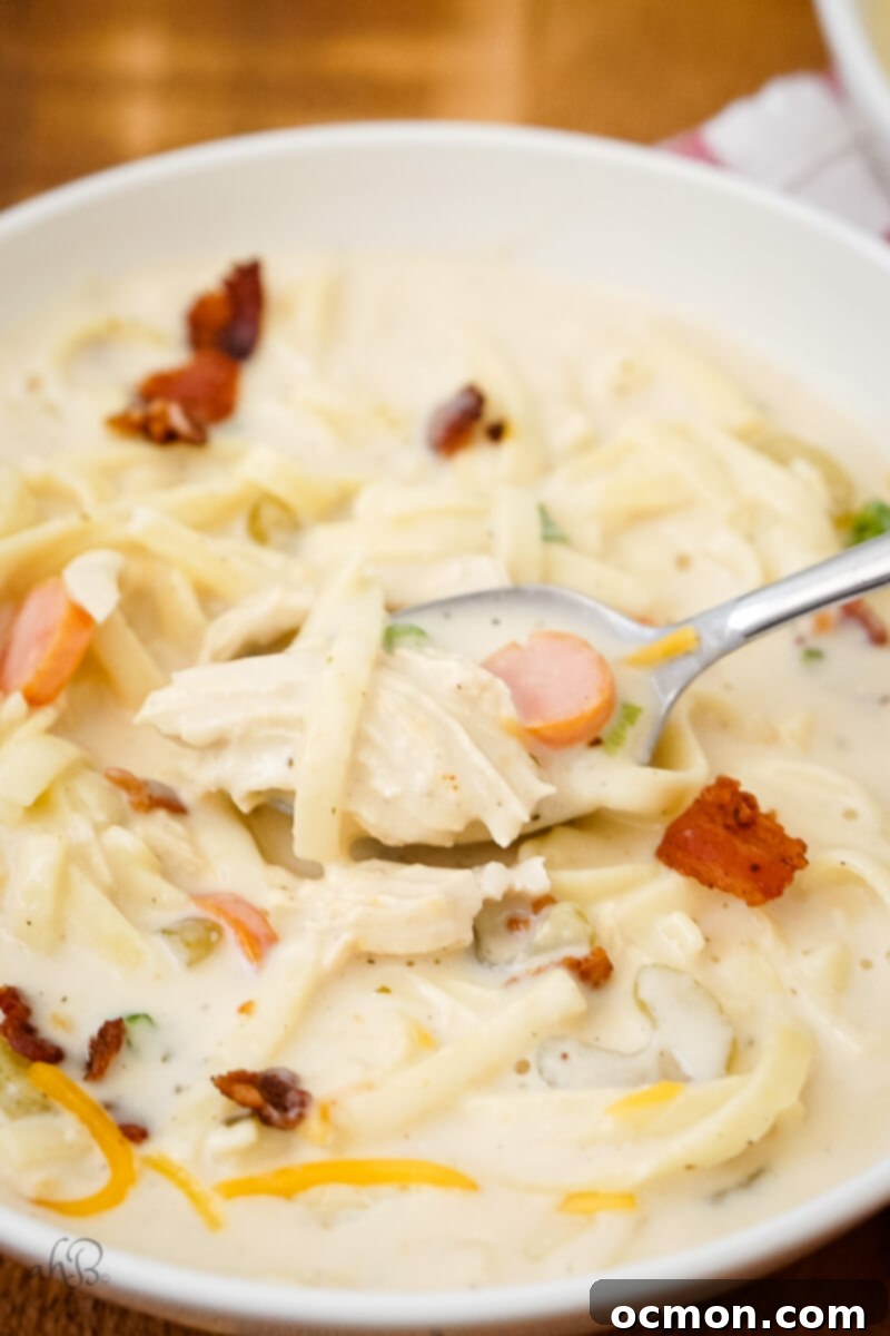 A close-up shot of a white bowl of Crack Chicken Noodle Soup, with a spoon scooping out a generous portion of noodles, chicken, and creamy broth, garnished with bacon and chives, highlighting its rich texture.