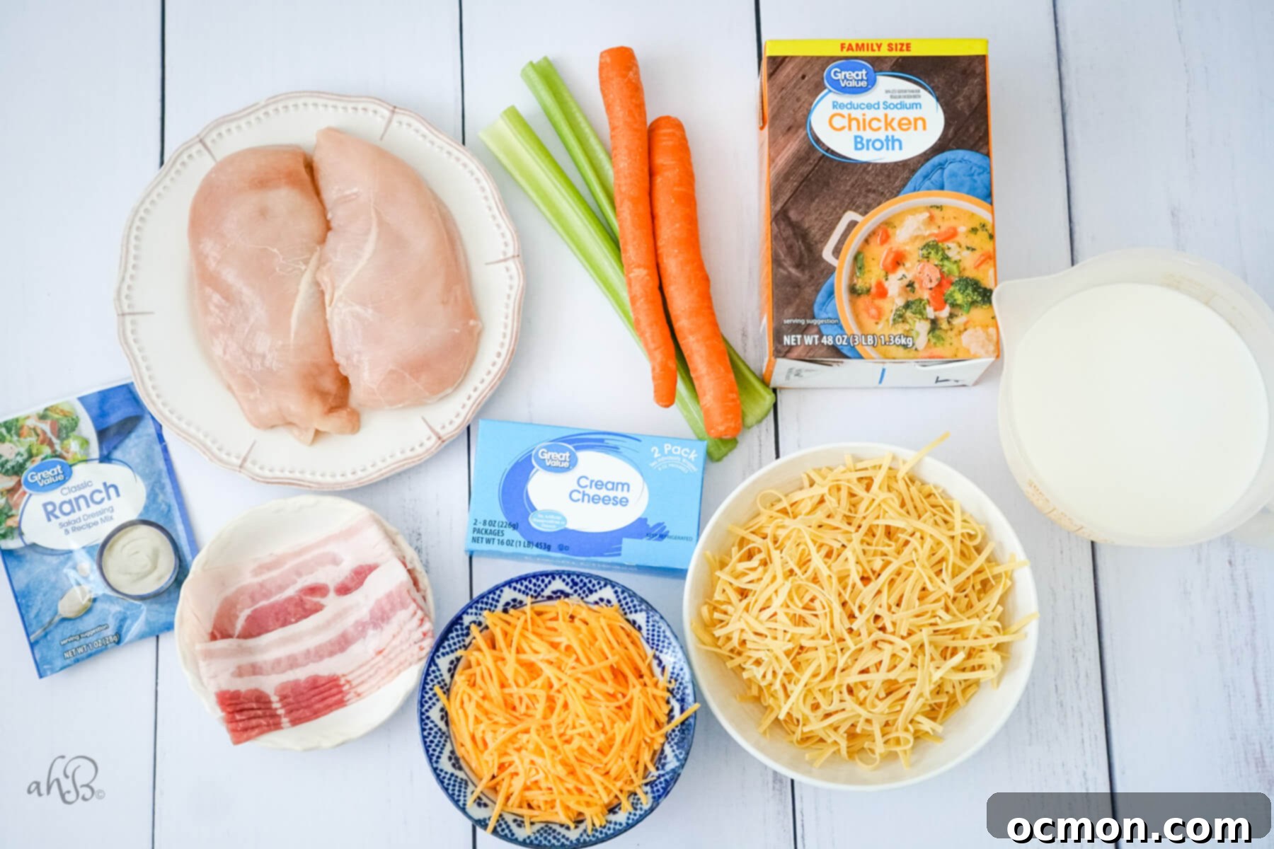 A flat lay of all the ingredients needed for Crack Chicken Noodle Soup: a ranch seasoning packet, crispy bacon strips, two raw chicken breasts, two carrots, two celery stalks, a block of cream cheese, shredded cheddar cheese, a bowl of dry egg noodles, a carton of milk, and a box of chicken broth, neatly arranged for preparation.