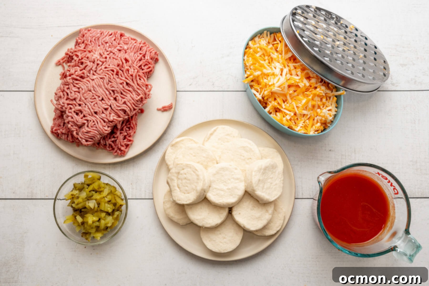 A flat lay of the 5 key ingredients for Sloppy Joe Biscuit Casserole: fresh ground beef, a can of sloppy joe sauce, chopped dill pickles, a can of refrigerated biscuits, and shredded Colby Jack cheese, neatly arranged on a white surface.