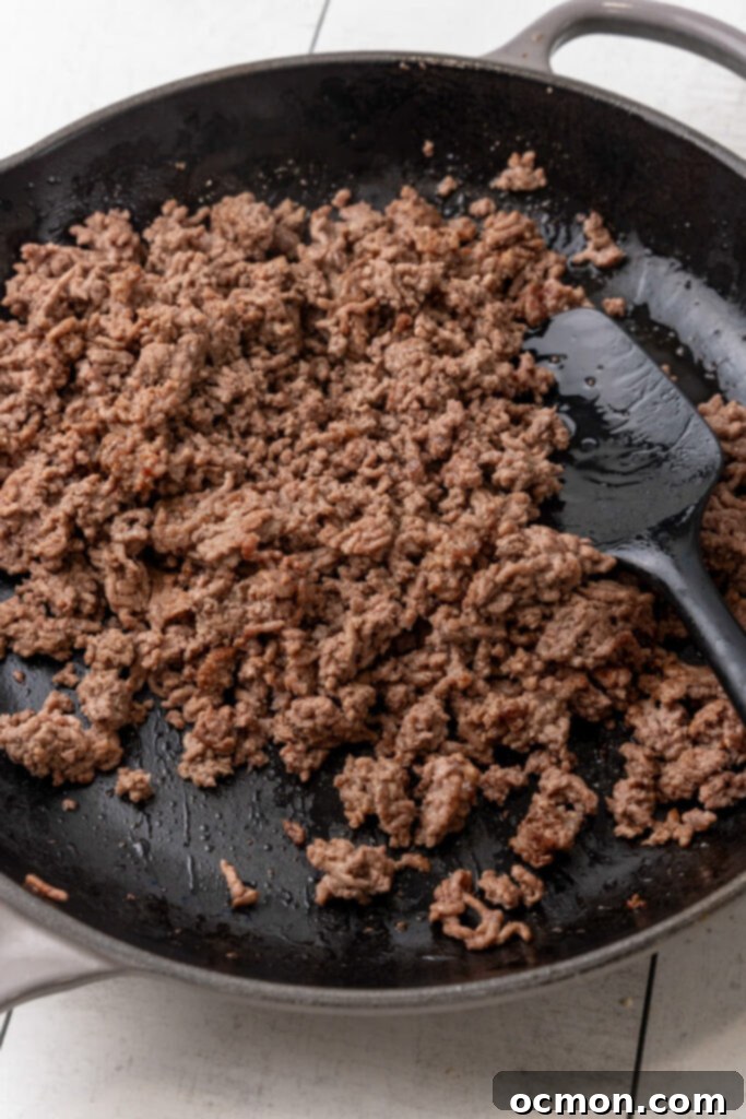 Ground beef browning in a large stainless steel skillet on the stovetop, being broken up with a wooden spoon.