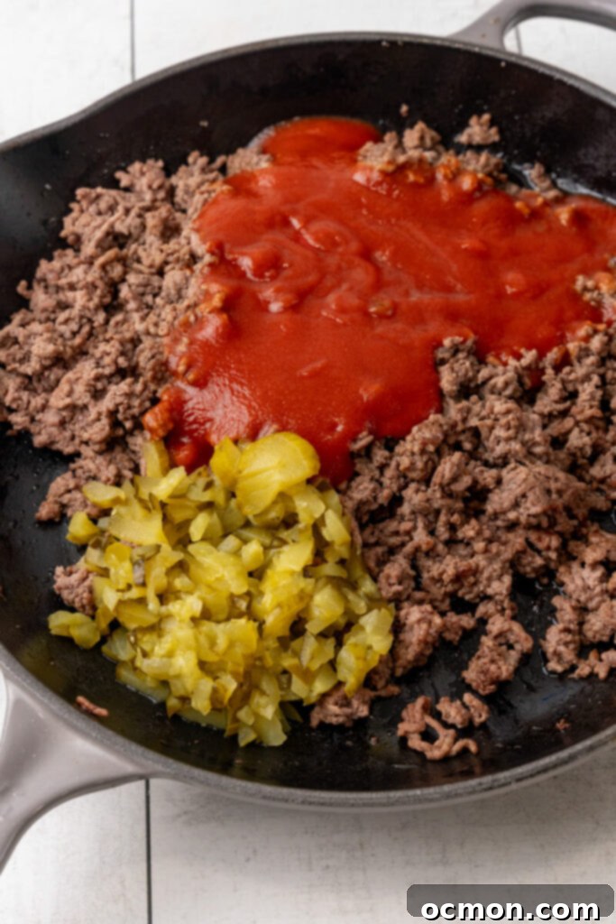 Sloppy joe sauce and chopped dill pickles being added to the browned ground beef in a skillet, ready to be stirred.