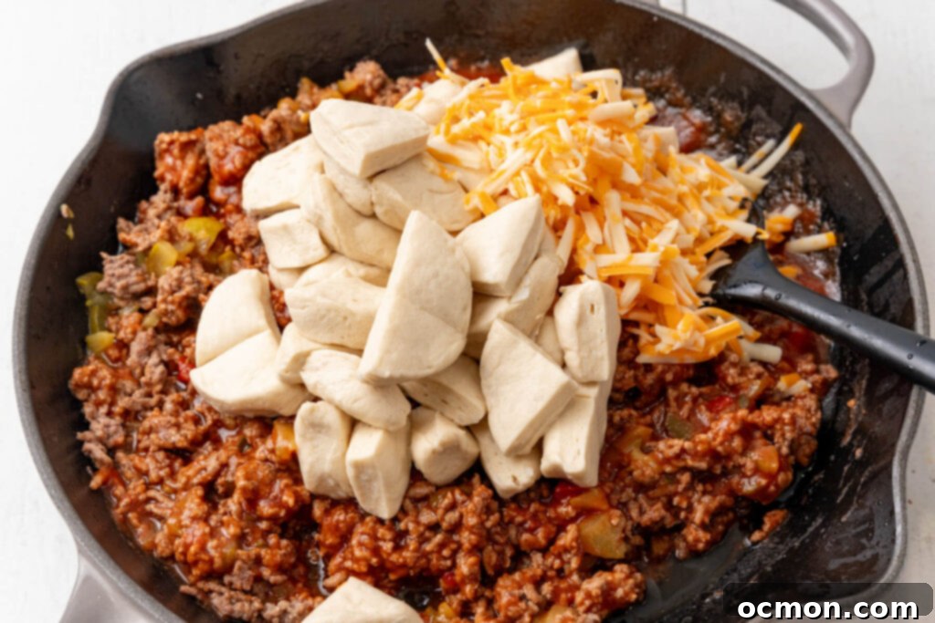 Biscuit pieces and shredded cheese being stirred into the sloppy joe meat mixture in a large skillet, before being transferred to a casserole dish.