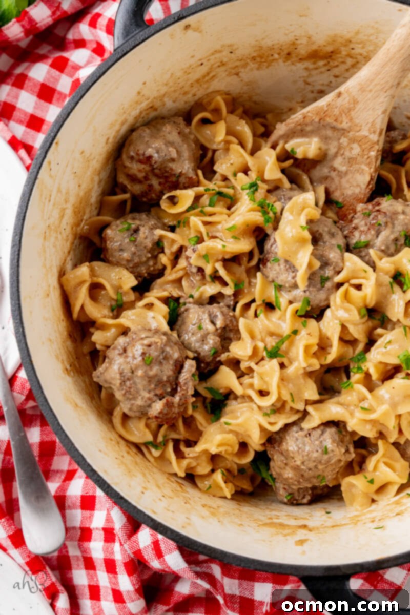 A pot of finished Swedish Meatballs stirred with a wooden spoon, highlighting the rich, creamy texture of the sauce and the tender noodles.